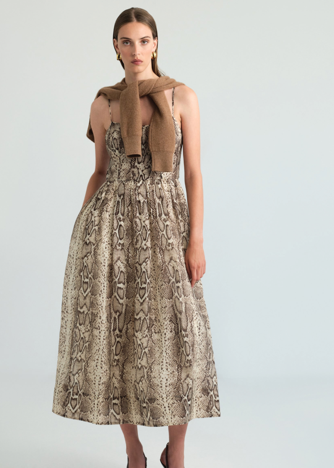 A model stands against a light gray studio background wearing a sleeveless midi dress with a beige and brown snakeskin print, a tan knit scarf tied around her shoulders, and dark brown heels.
