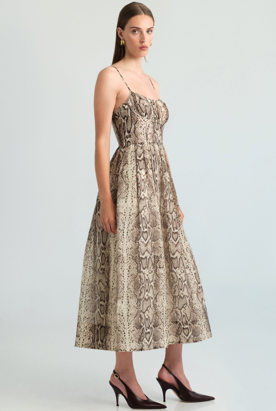 A model stands against a light gray studio background wearing a sleeveless midi dress with a beige and brown snakeskin print, a tan knit scarf tied around her shoulders, and dark brown heels.