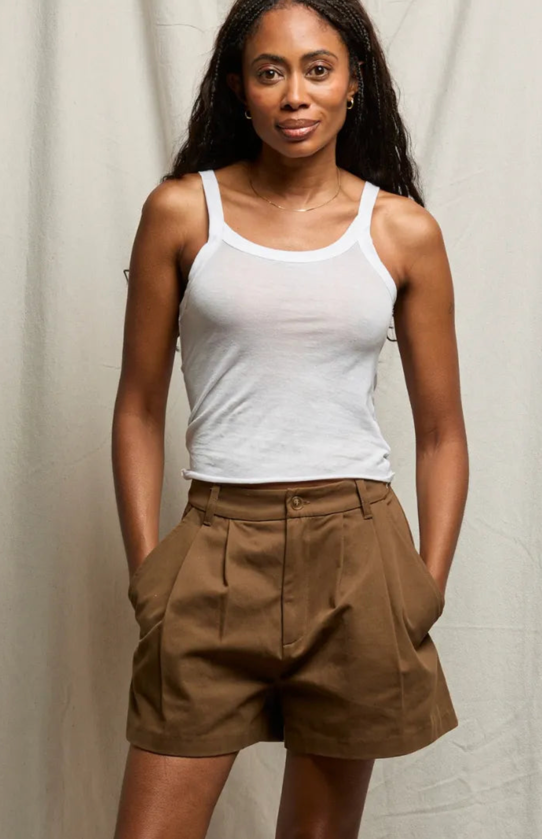 A woman wearing a sheer black tank top and light beige trousers, posing against a light gray fabric backdrop.