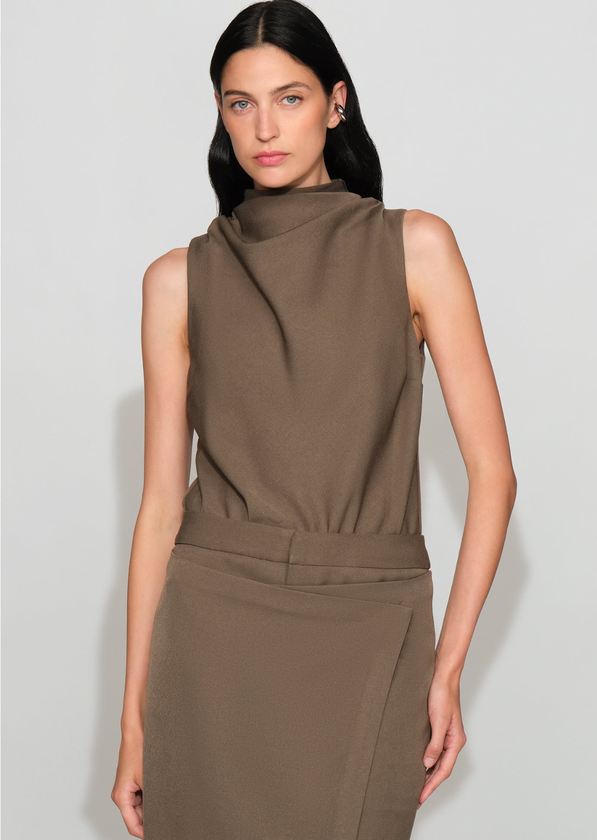 A model wearing a sleeveless, high-neck, draped brown dress against a light gray studio background.