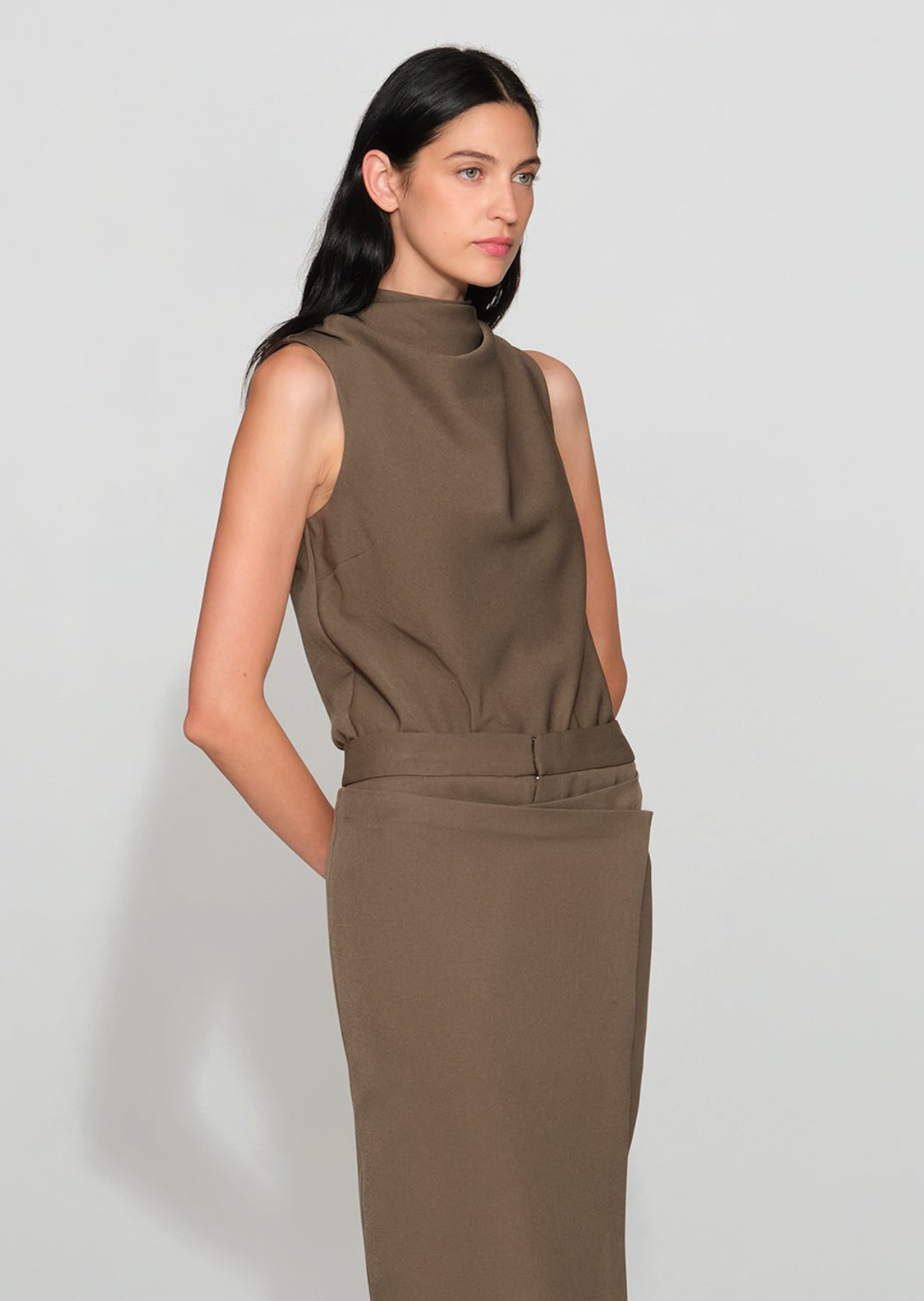A model wearing a sleeveless, high-neck, draped brown dress against a light gray studio background.