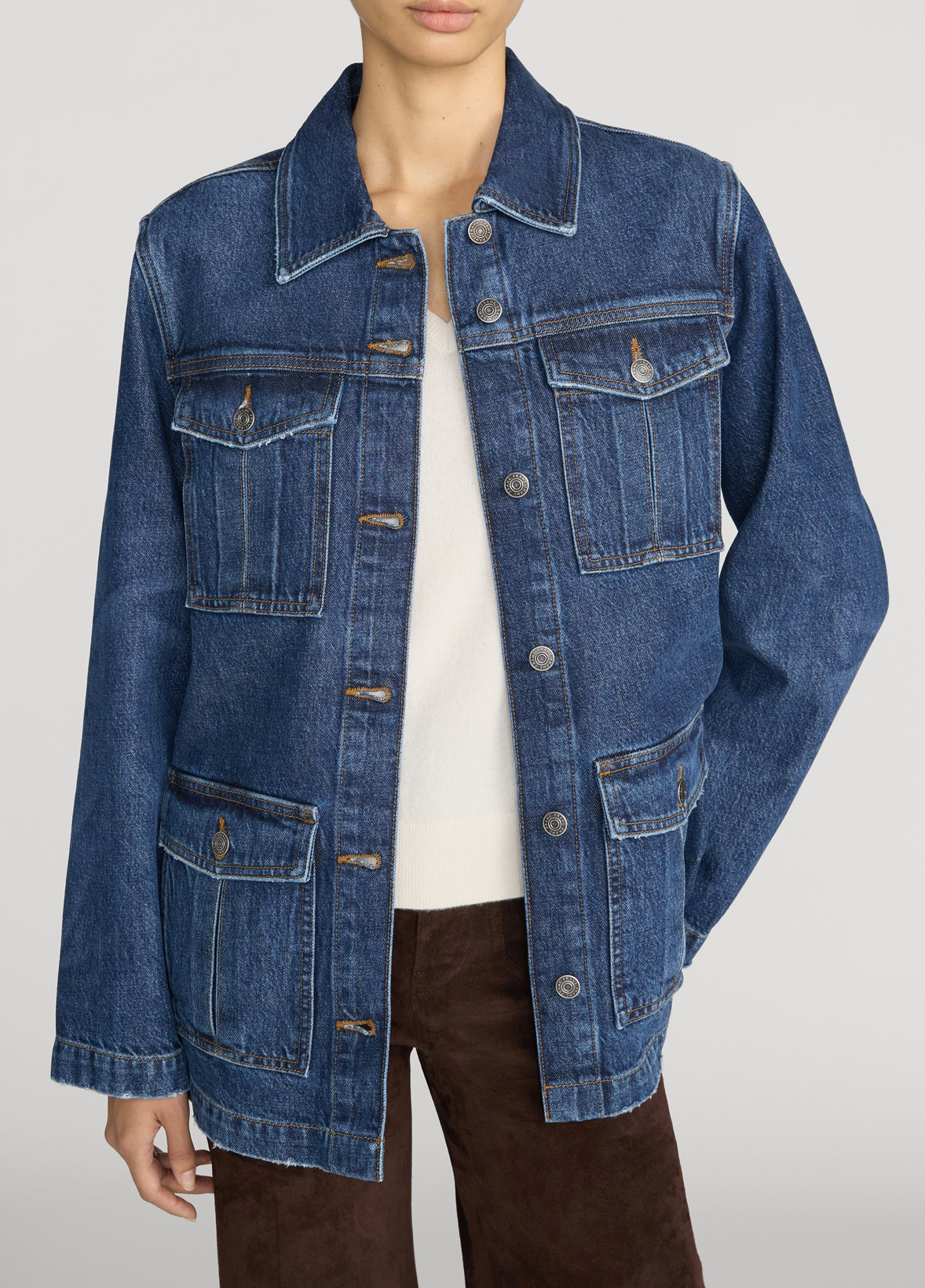 A model wearing a blue denim trucker jacket over a white top and brown pants, shown from the neck down against a light gray studio background.