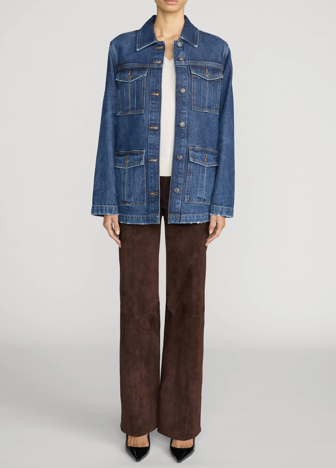 A model wearing a blue denim trucker jacket over a white top and brown pants, shown from the neck down against a light gray studio background.