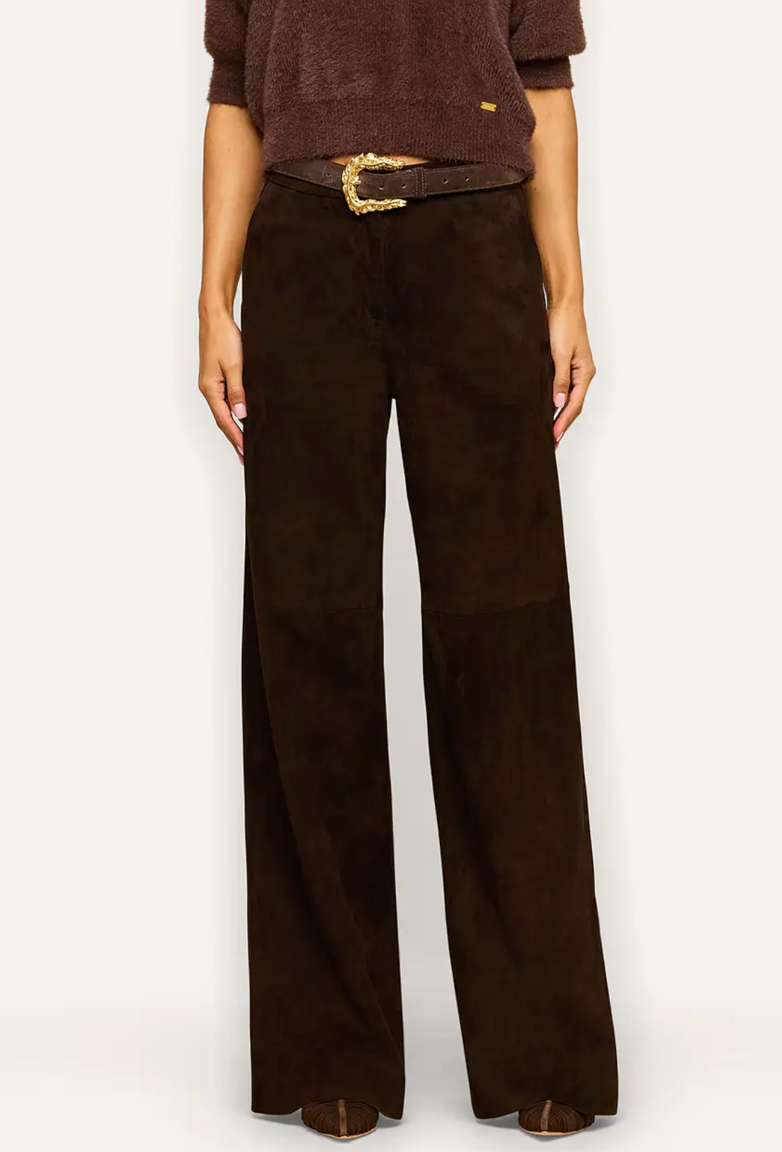 A woman modeling a dark brown short-sleeve knit top and dark brown wide-leg trousers with a gold-buckled belt, photographed against a plain light gray studio background.