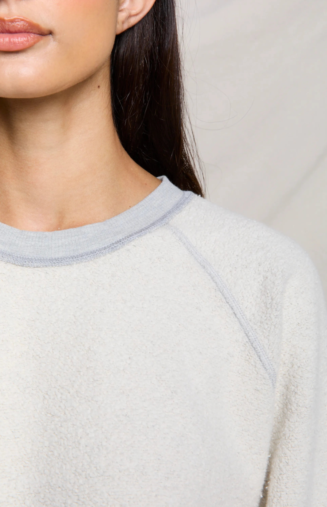 A woman wearing a cream-colored, textured fleece crewneck sweatshirt with light gray trim on the collar and cuffs, paired with blue jeans, standing against a draped beige fabric backdrop.