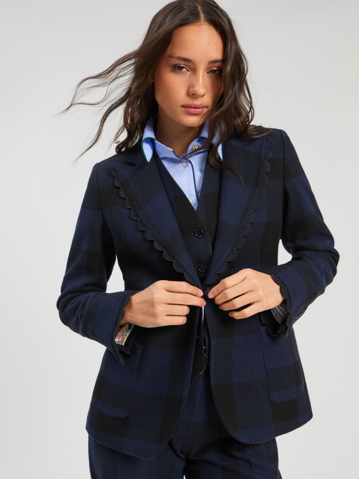 A woman wearing a dark navy and black plaid blazer with scalloped lapels, styled over a matching vest and a light blue shirt, against a plain light gray studio background.