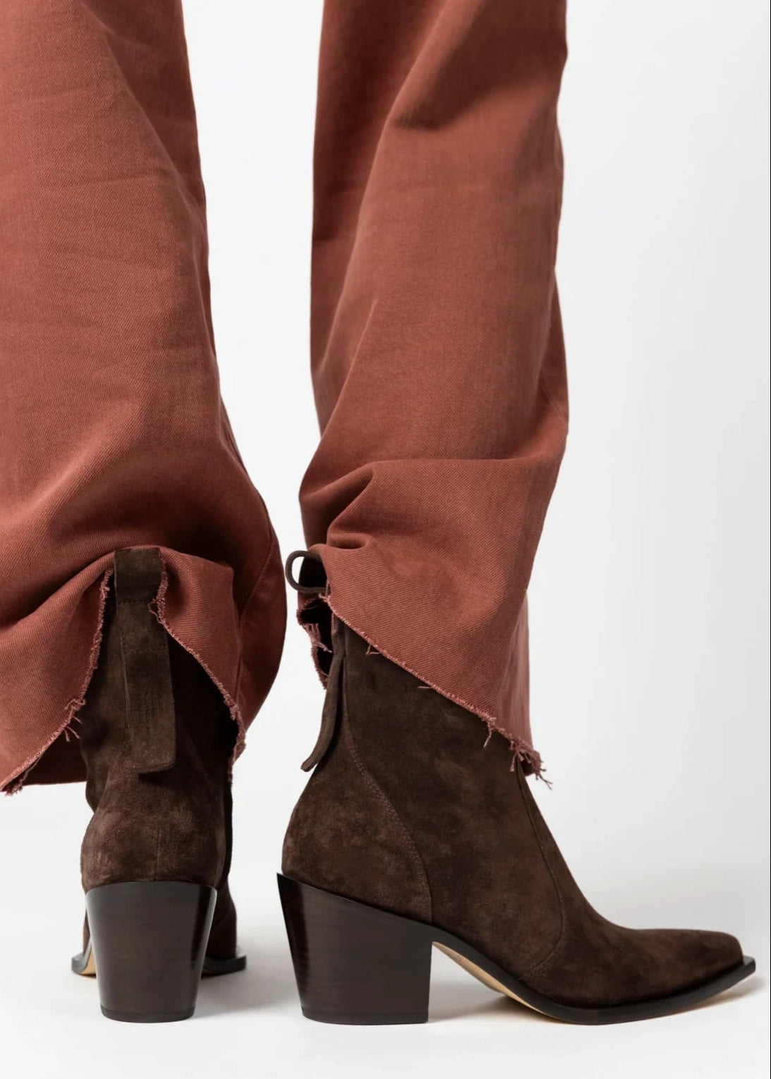 A close-up of a person's legs wearing brown suede ankle boots with block heels and pointed toes, paired with rust-colored pants against a white background.