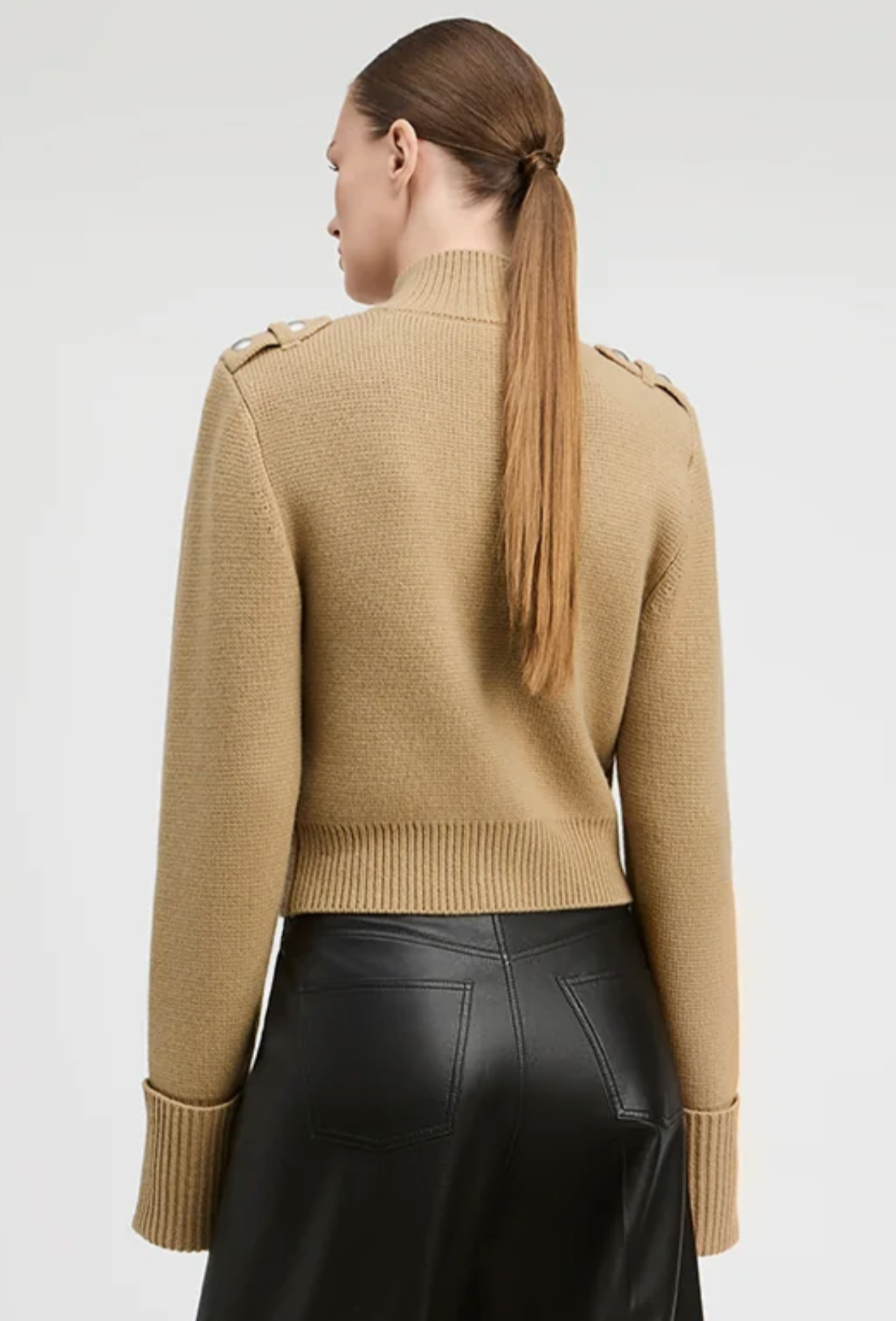 A woman shown from the back wearing a camel beige knit mock-neck sweater with shoulder button details and black leather pants, against a light gray studio background.