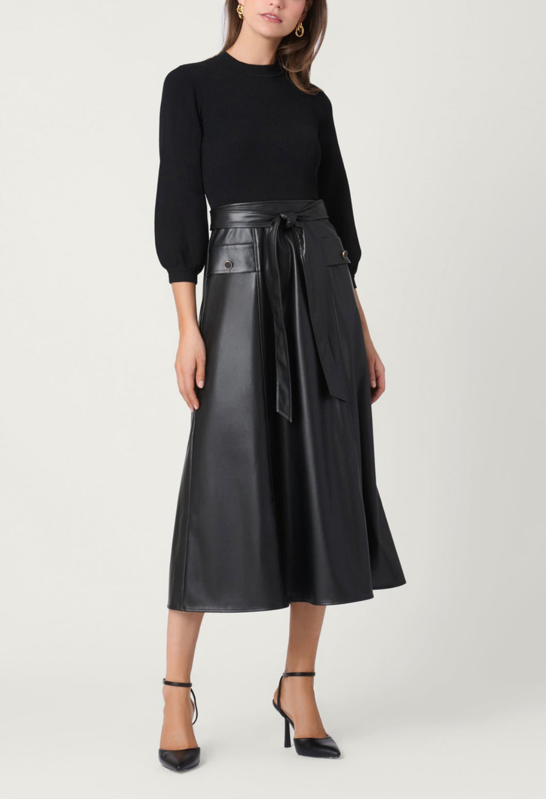 A model wearing a black long-sleeve knit top and a black faux-leather midi skirt with a tie waist, paired with black high-heeled sandals, against a light gray studio background.