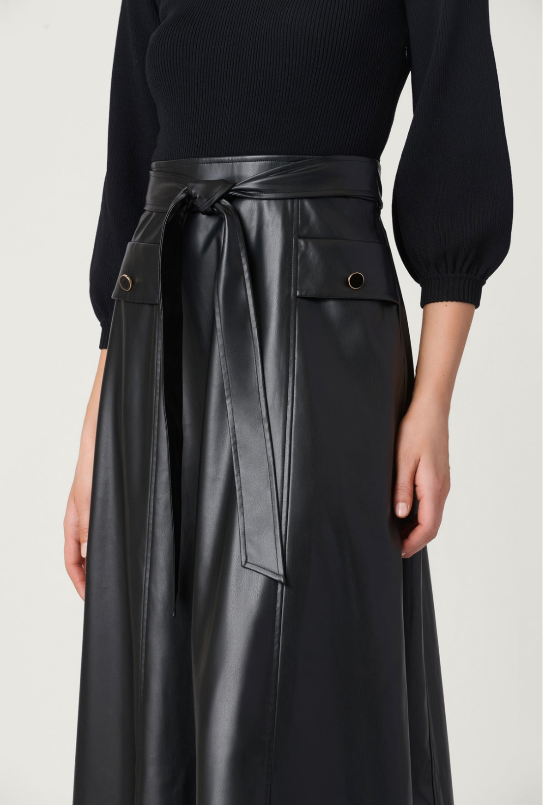 A model wearing a black long-sleeve knit top and a black faux-leather midi skirt with a tie waist, paired with black high-heeled sandals, against a light gray studio background.