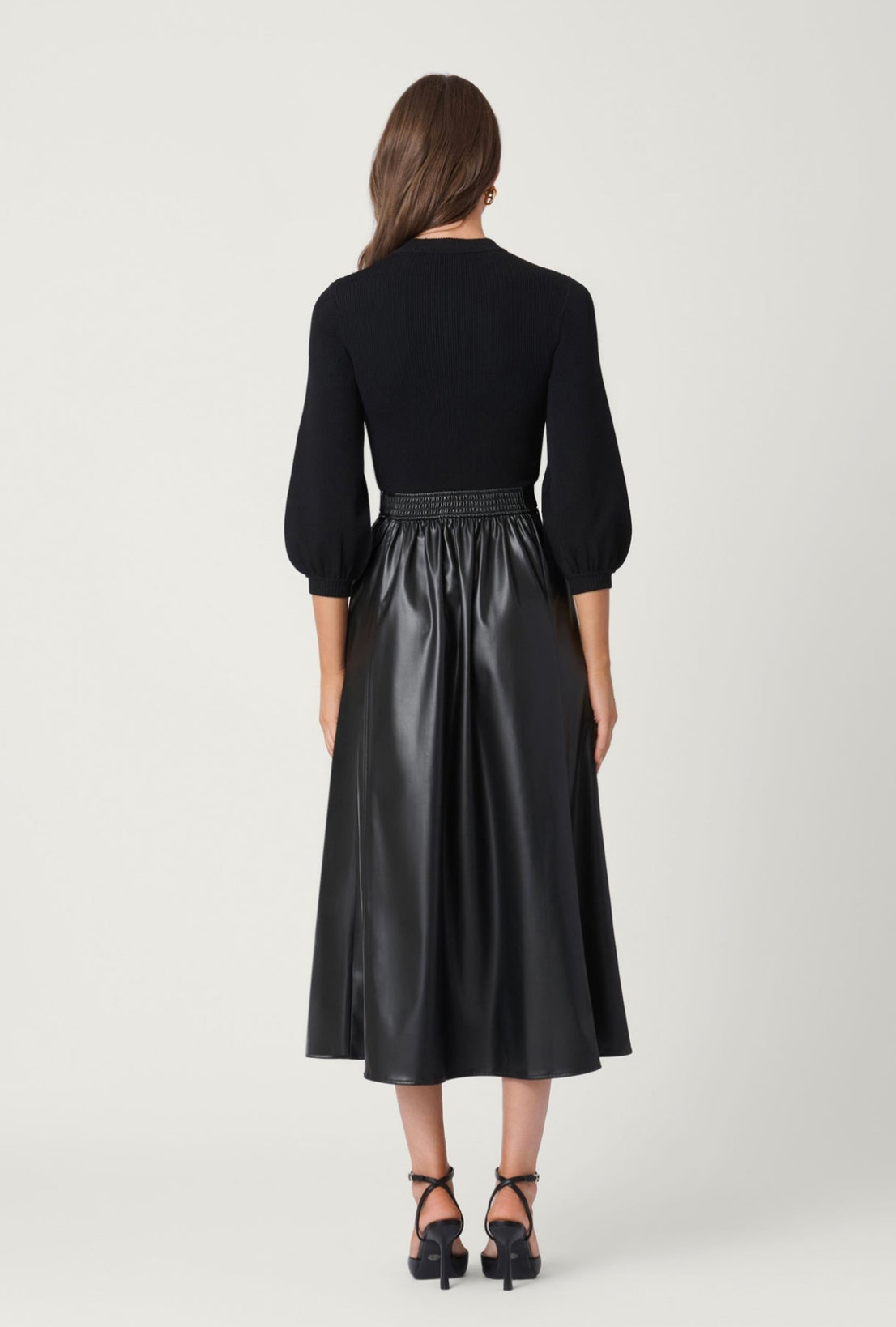 A woman viewed from the back wearing a black long-sleeve top with puff sleeves and a black midi skirt with a shiny finish and a textured waistband, paired with black heels, against a light gray studio background.