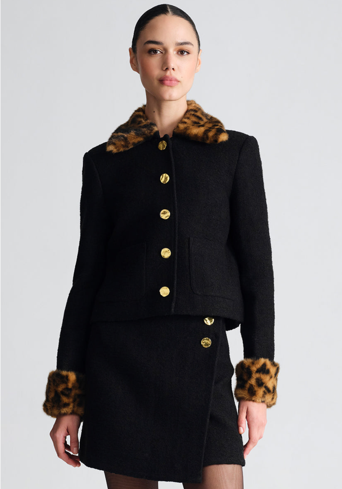 A model wears a black tweed jacket and matching skirt set featuring leopard-print fur trim on the collar and cuffs, accented with large gold buttons, set against a plain light gray studio background.