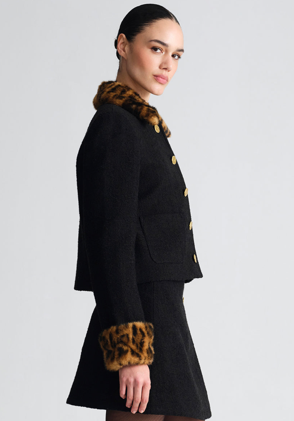 A model wears a black tweed jacket and matching skirt set featuring leopard-print fur trim on the collar and cuffs, accented with large gold buttons, set against a plain light gray studio background.