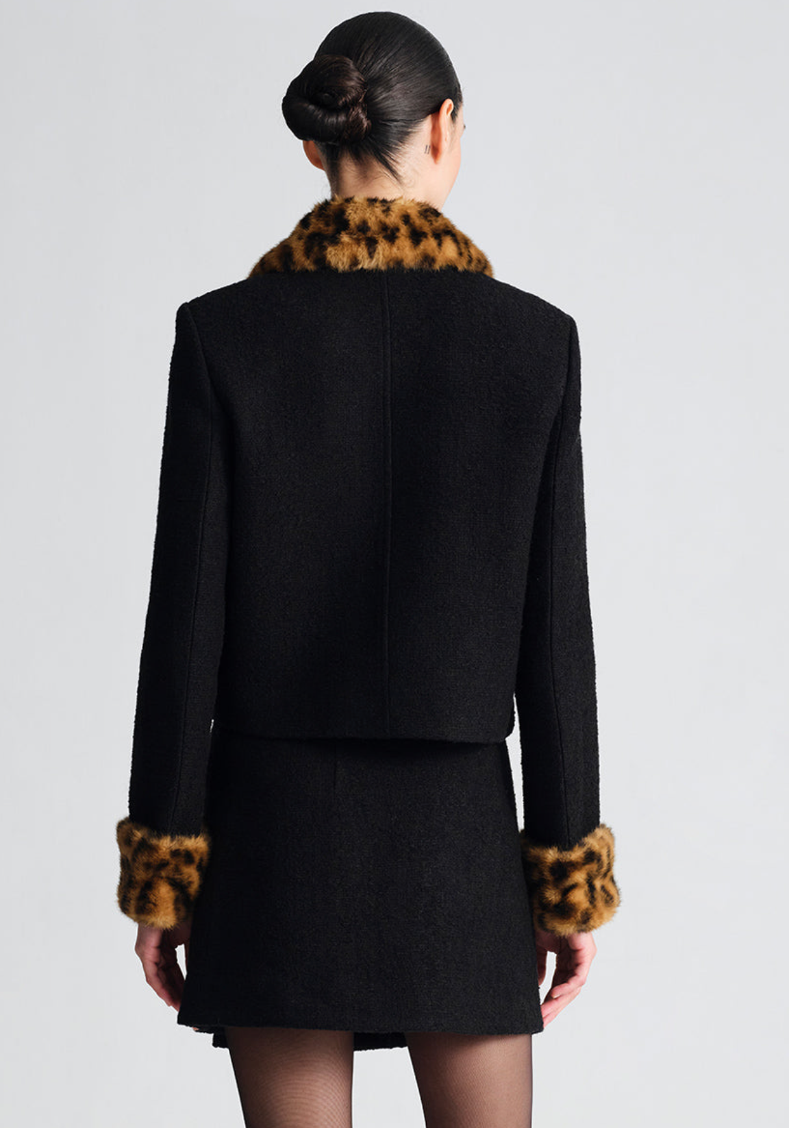 A model shown from the back wearing a black cropped jacket and matching mini skirt with brown and black leopard-print fur trim at the collar and cuffs, set against a plain light gray studio background.
