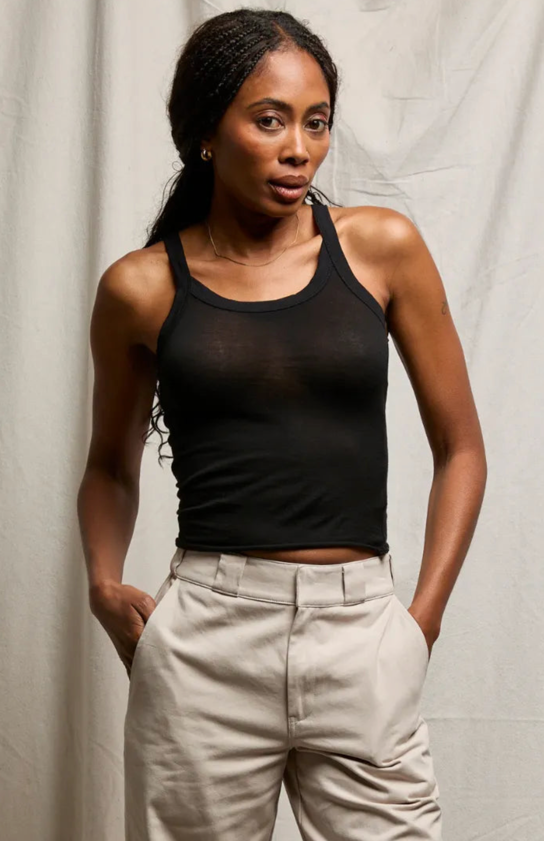A woman wearing a sheer black tank top and light beige trousers, posing against a light gray fabric backdrop.