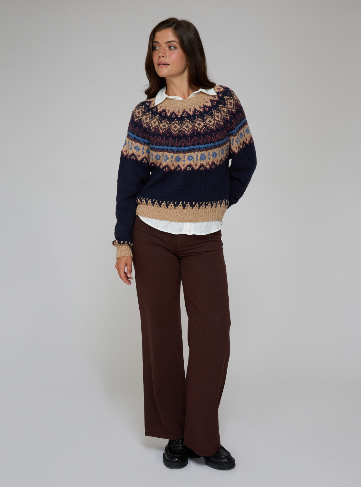 A woman wearing a navy blue knit sweater with beige and light blue Fair Isle patterns, layered over a white collared shirt and black pants, standing in a subway station.