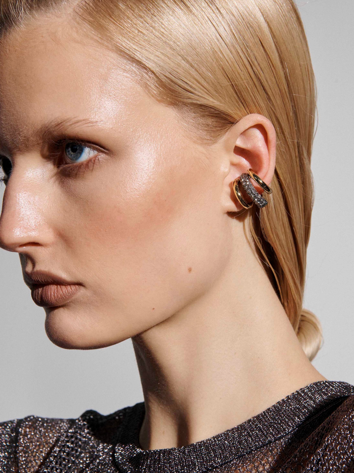 Close-up of a blonde model wearing a gold hoop earring with a textured, dark, rhinestone-like center, set against a neutral gray background.