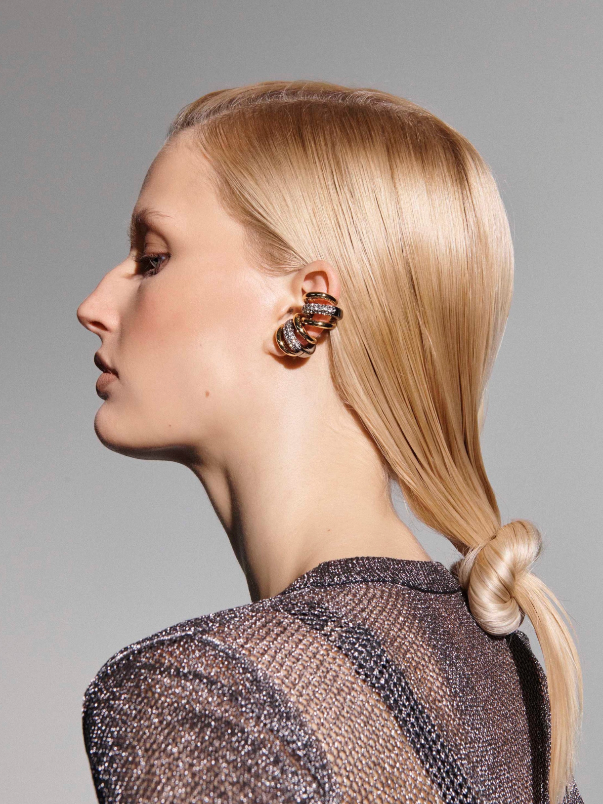 A side-profile portrait of a blonde woman wearing a chunky, multi-layered spiral earring with metallic gold and silver-toned bands set with clear stones, paired with a sparkly dark top against a neutral gray background.