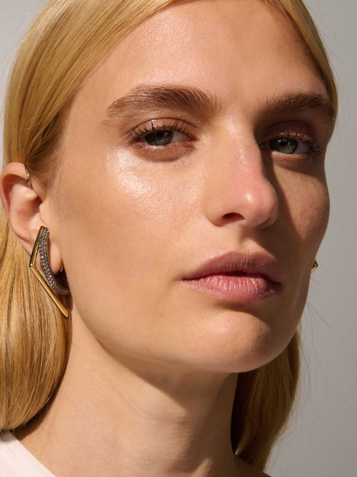 Close-up of a blonde woman wearing a gold geometric hoop earring with pavé-set clear stones, set against a neutral gray background.