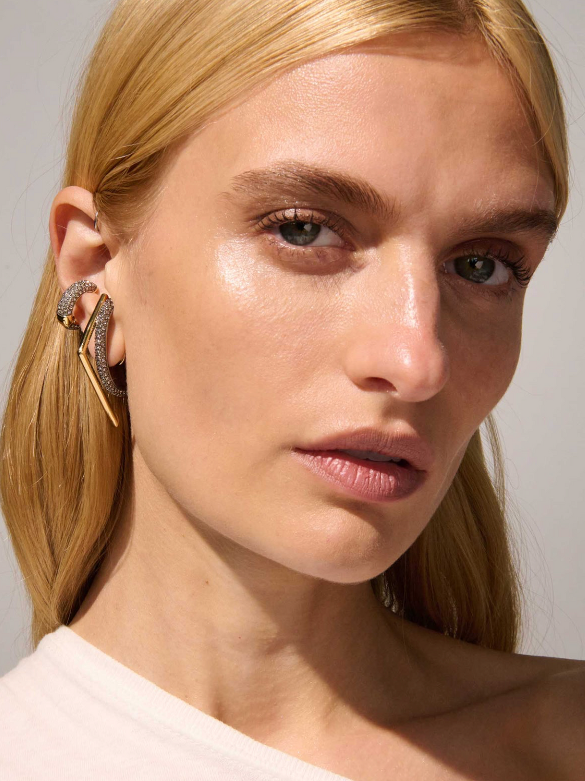 A close-up of a blonde woman wearing a pair of textured gold hoop earrings with small red and green gemstone accents, set against a plain light gray background.
