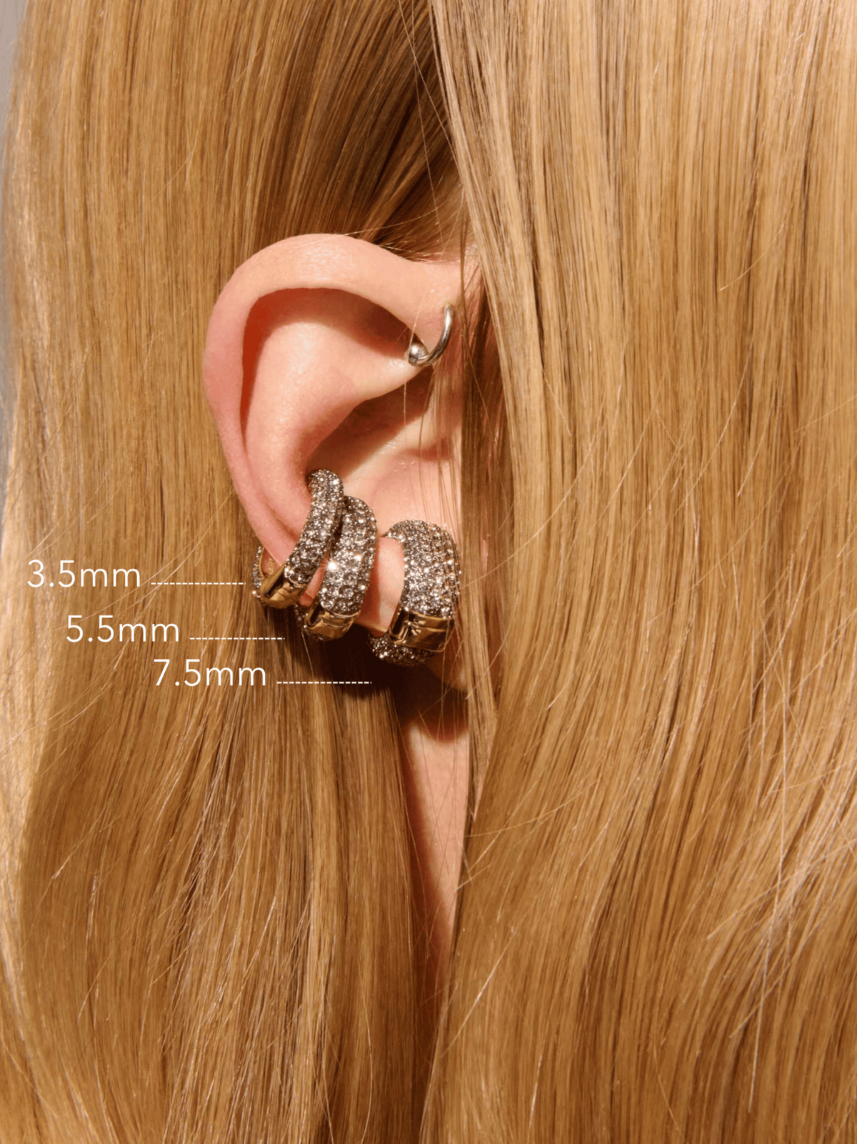 Close-up of a person's ear wearing three silver hoop earrings with clear stones, with size labels showing 3.5mm, 5.
