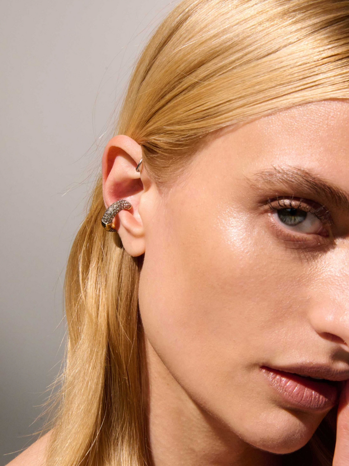 Close-up of a blonde woman's ear wearing a textured silver hoop earring and a small silver cartilage piercing against a neutral gray background.