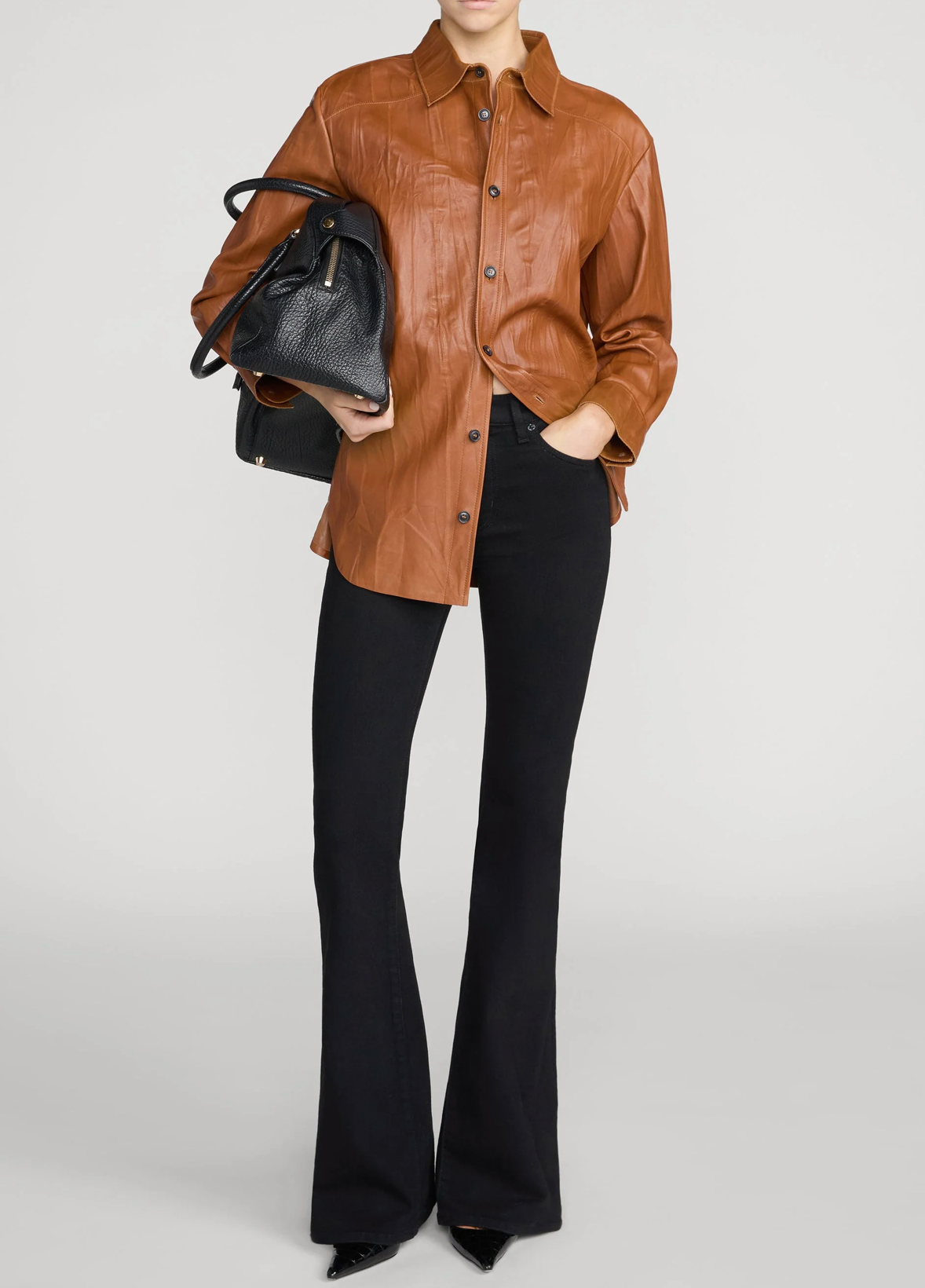 A model wearing a brown leather button-up shirt, black flared pants, and holding a black textured handbag against a light gray studio background.