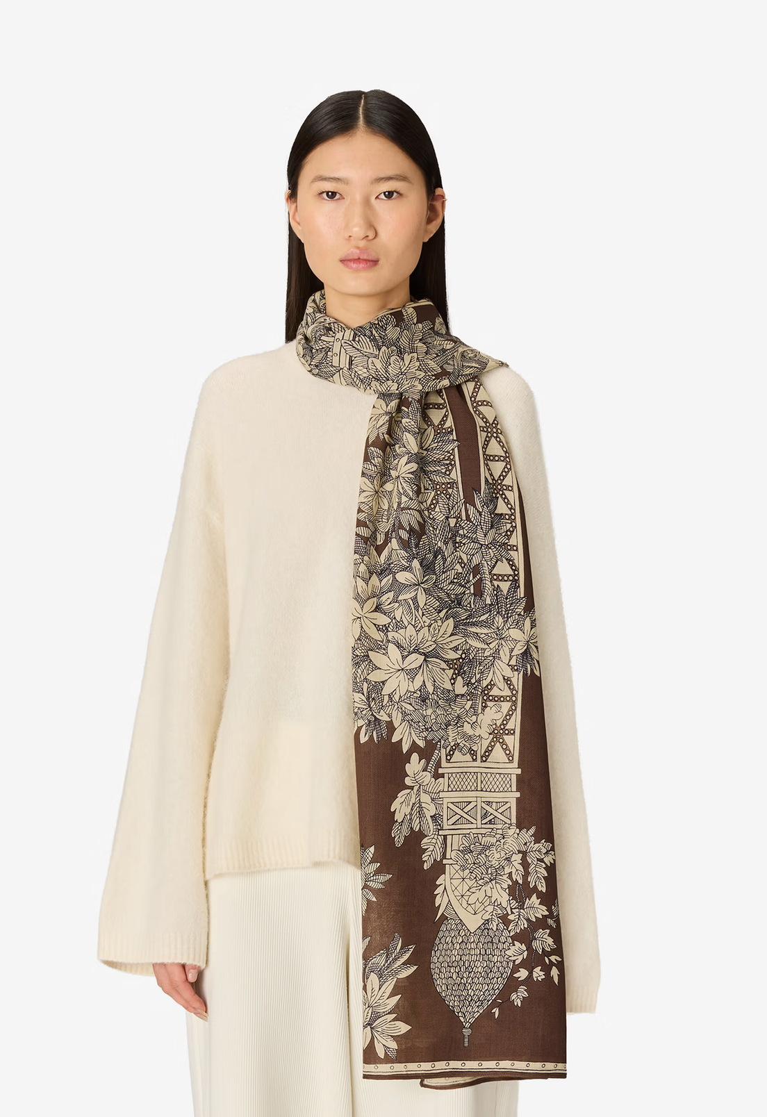 A model wearing a cream knit sweater and white trousers, styled with a brown and beige patterned silk scarf featuring botanical and architectural motifs, set against a plain light gray background.