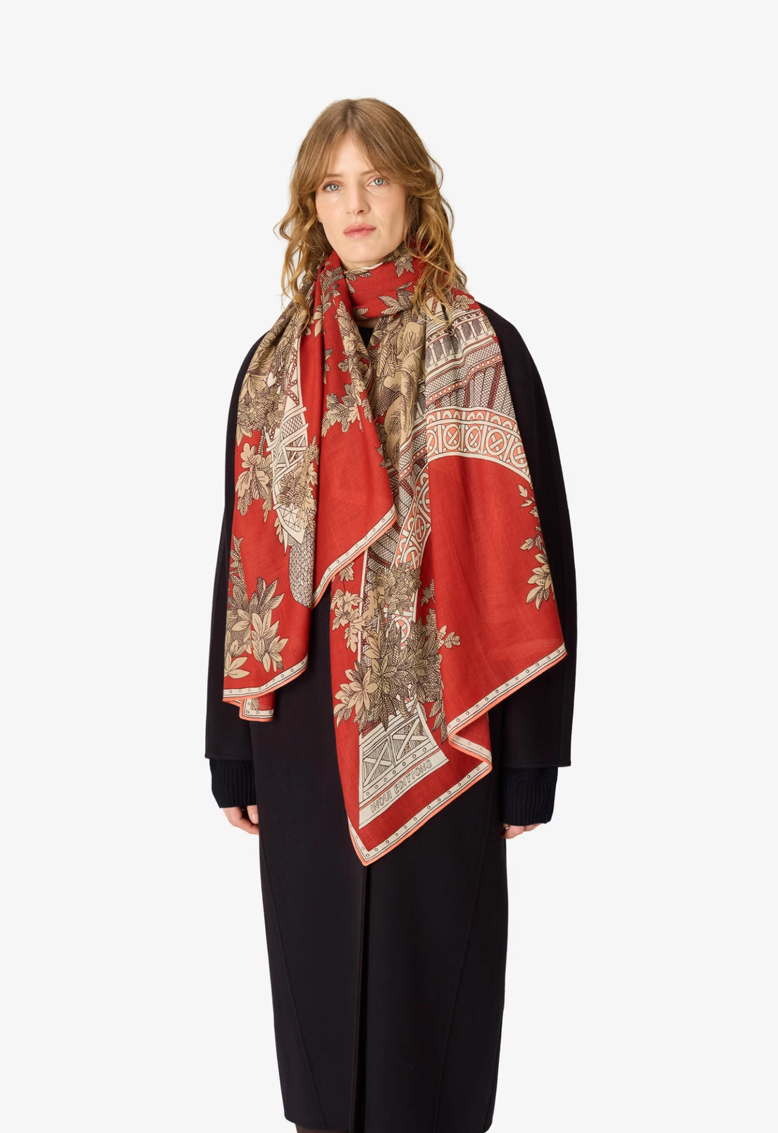 A model wearing a long red silk scarf with a detailed beige and brown botanical and architectural pattern, styled over a black coat against a plain white background.