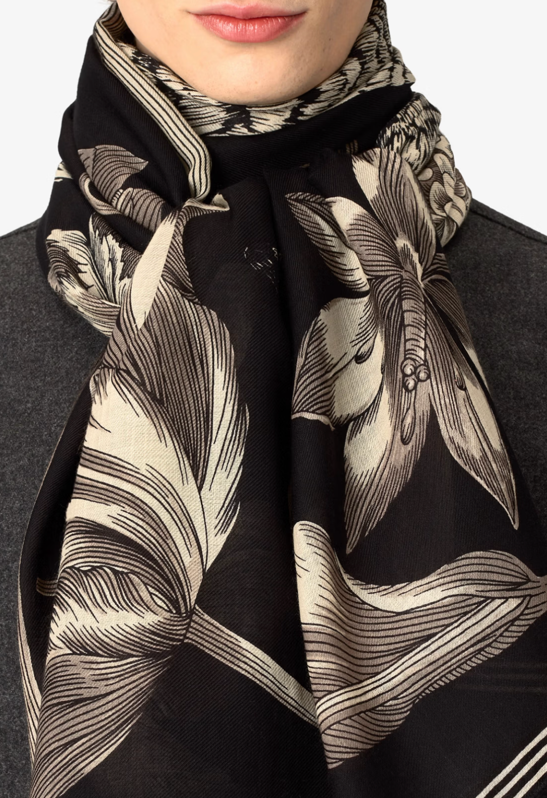 A close-up of a model wearing a black scarf with a large beige botanical line-art print, styled over a dark gray coat against a light gray background.