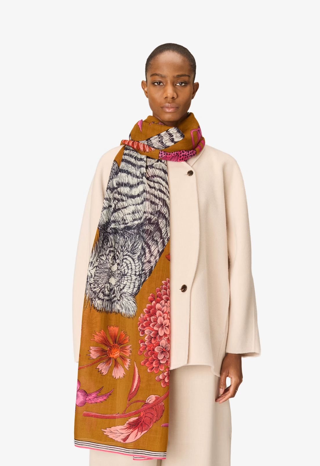 A model wearing a cream-colored coat and trousers, styled with a mustard yellow silk scarf featuring a tiger and floral print, against a plain white background.