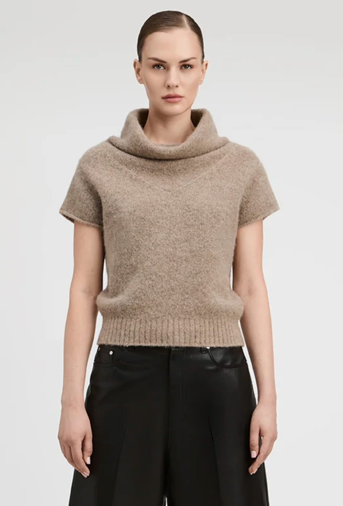 A model wearing a beige short-sleeve cowl-neck knit sweater and black wide-leg trousers, photographed against a light gray studio background.