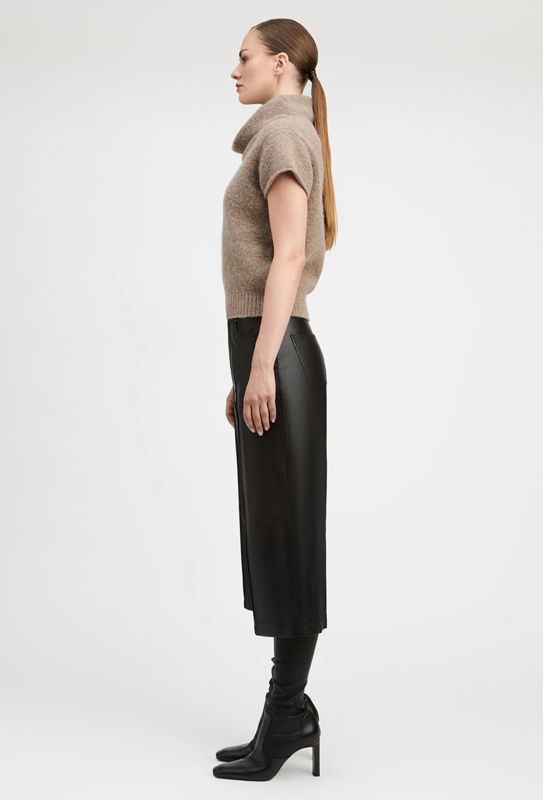 A woman in profile wearing a beige short-sleeve knit sweater, a black midi skirt, and black heeled ankle boots, photographed against a clean light gray studio background.