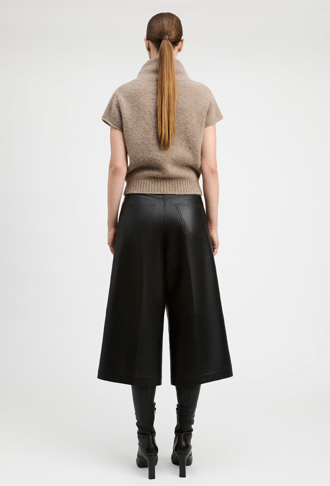 A model shown from the back wearing a beige short-sleeve knit mock-neck sweater, black wide-leg cropped pants, and black heeled boots against a light gray studio background.