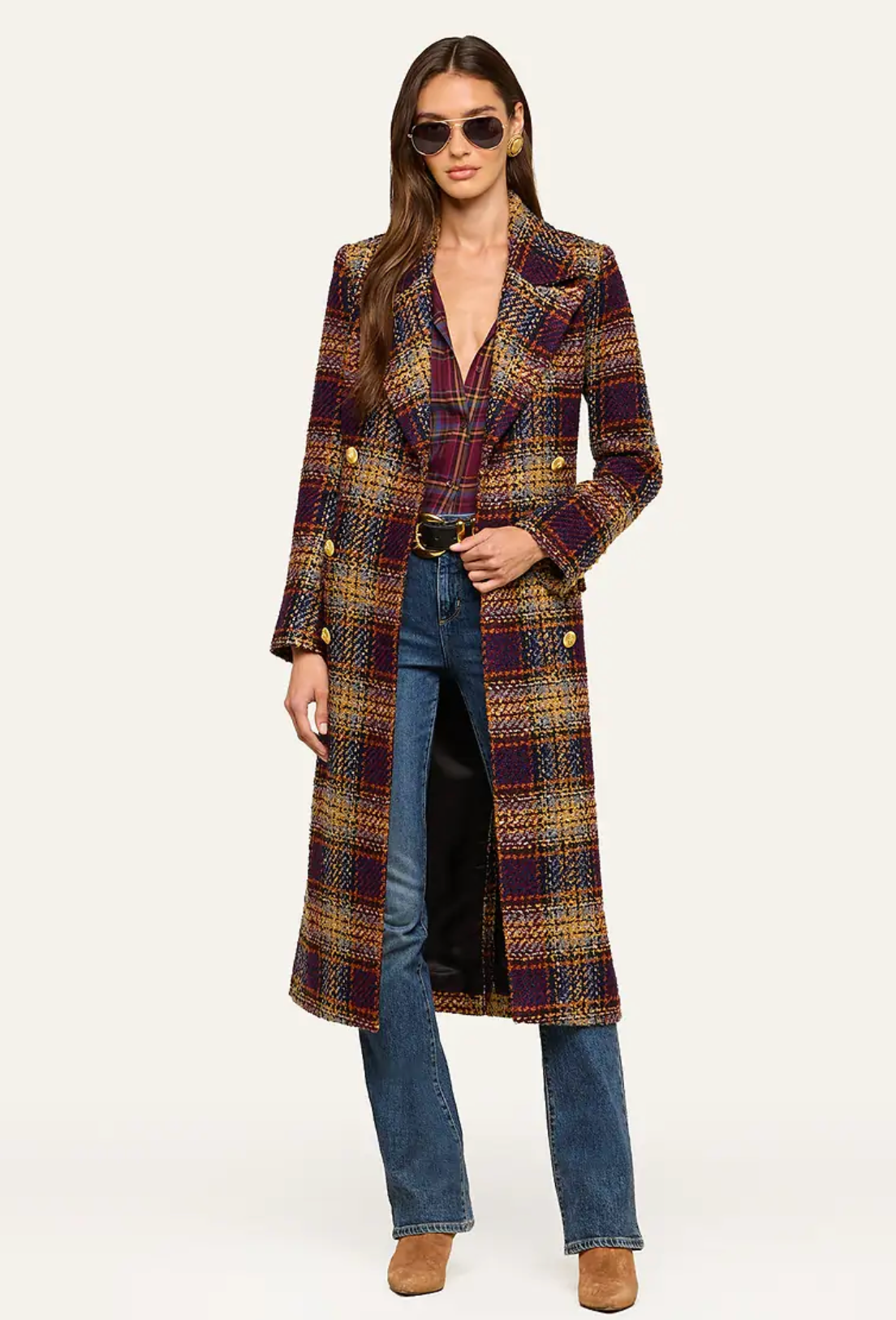 A model wearing a long, double-breasted plaid coat in purple, gold, and brown tones, styled with blue jeans, a black top, and brown suede boots against a plain white studio background.