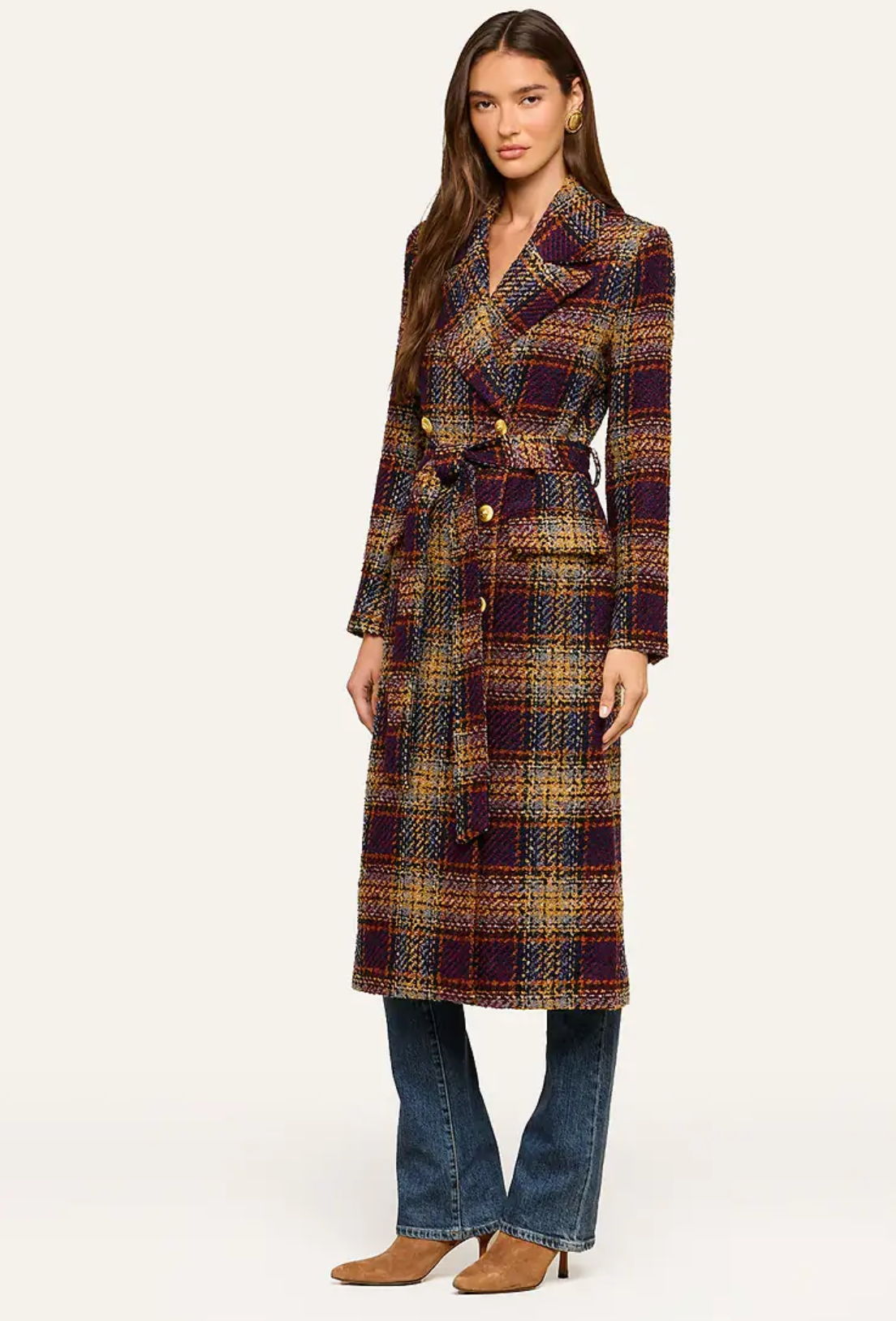 A woman modeling a long plaid coat with a waist tie, paired with blue jeans and brown ankle boots, against a plain light background.