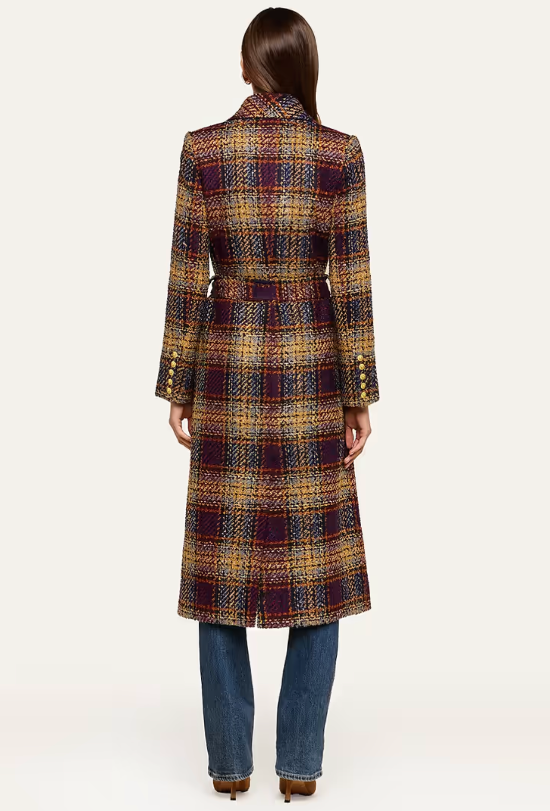 A woman viewed from the back wearing a long plaid coat in brown, gold, and dark purple tones, paired with blue jeans and brown heels, against a plain light background.