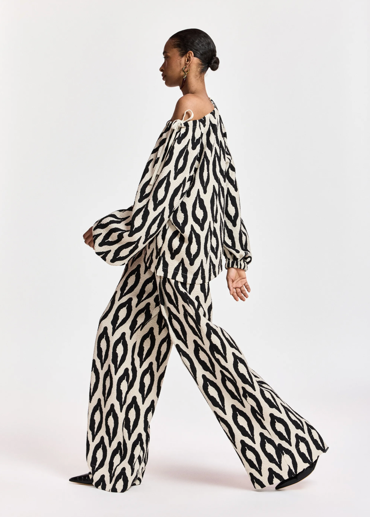 A model in profile wearing a matching off-the-shoulder long-sleeve top and wide-leg trousers set in a black and off-white ikat pattern, photographed against a seamless light gray studio background.