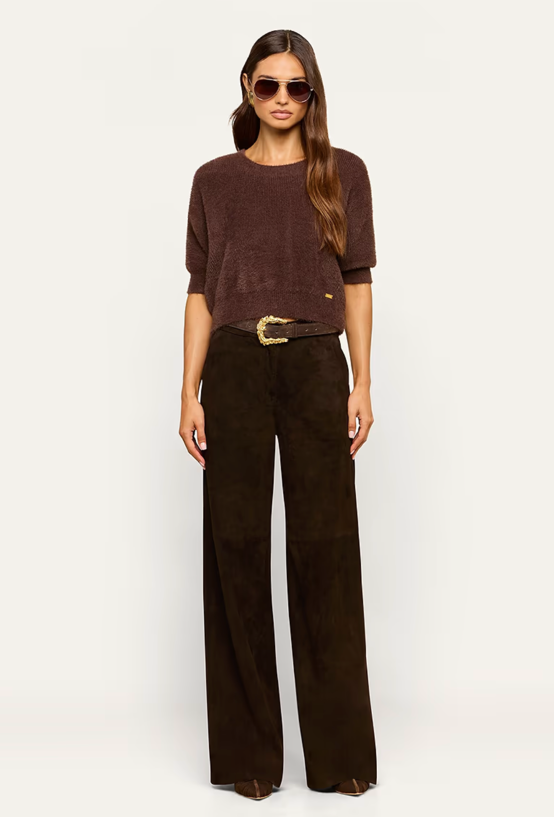A woman modeling a dark brown short-sleeve knit top and dark brown wide-leg trousers with a gold-buckled belt, photographed against a plain light gray studio background.