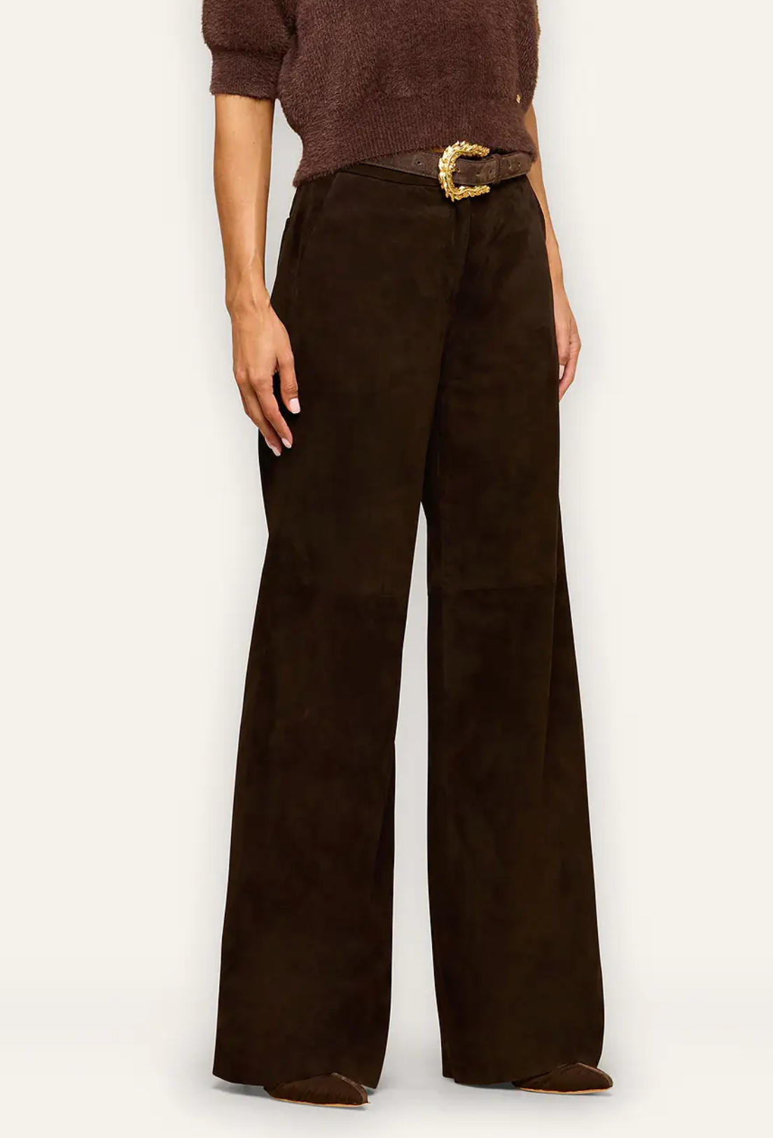 A model wearing dark brown wide-leg trousers made of a soft suede-like material, styled with a brown knit sweater, a brown belt with a gold buckle, and brown flat shoes against a plain off-white background.