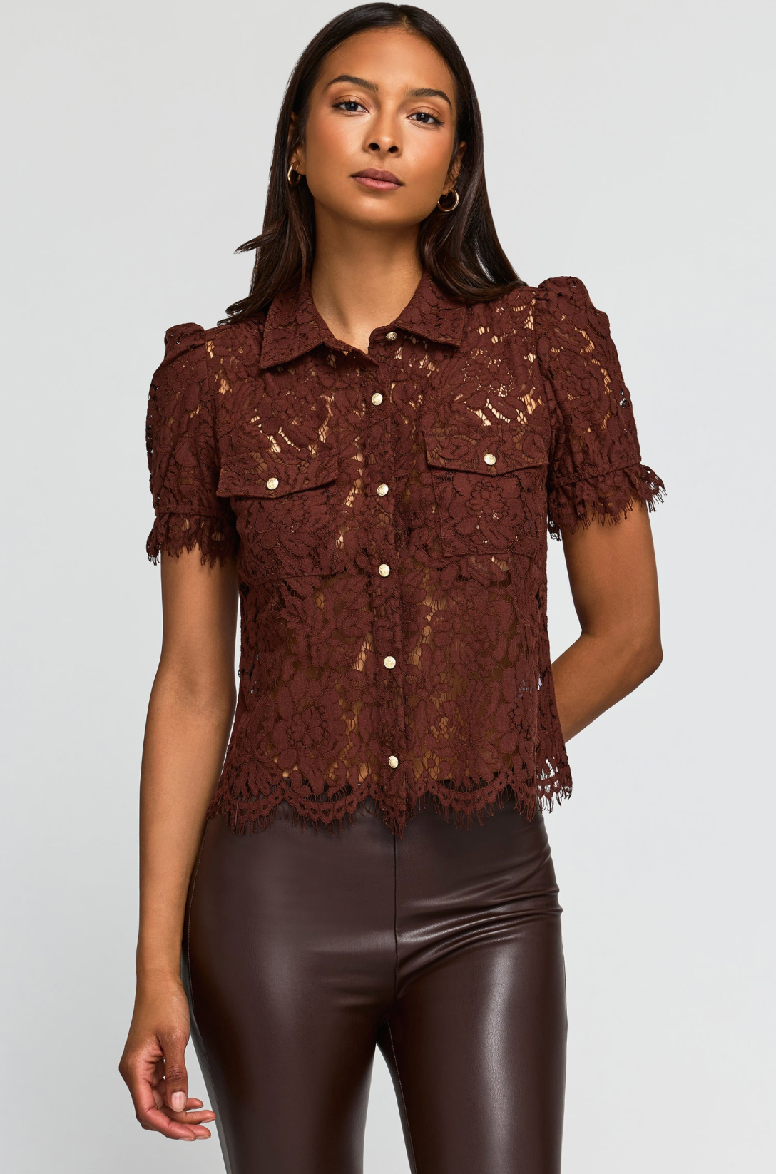 A woman wearing a short-sleeve lace button-up blouse in dark brown with matching brown pants, standing outdoors in front of a stone wall and green foliage.