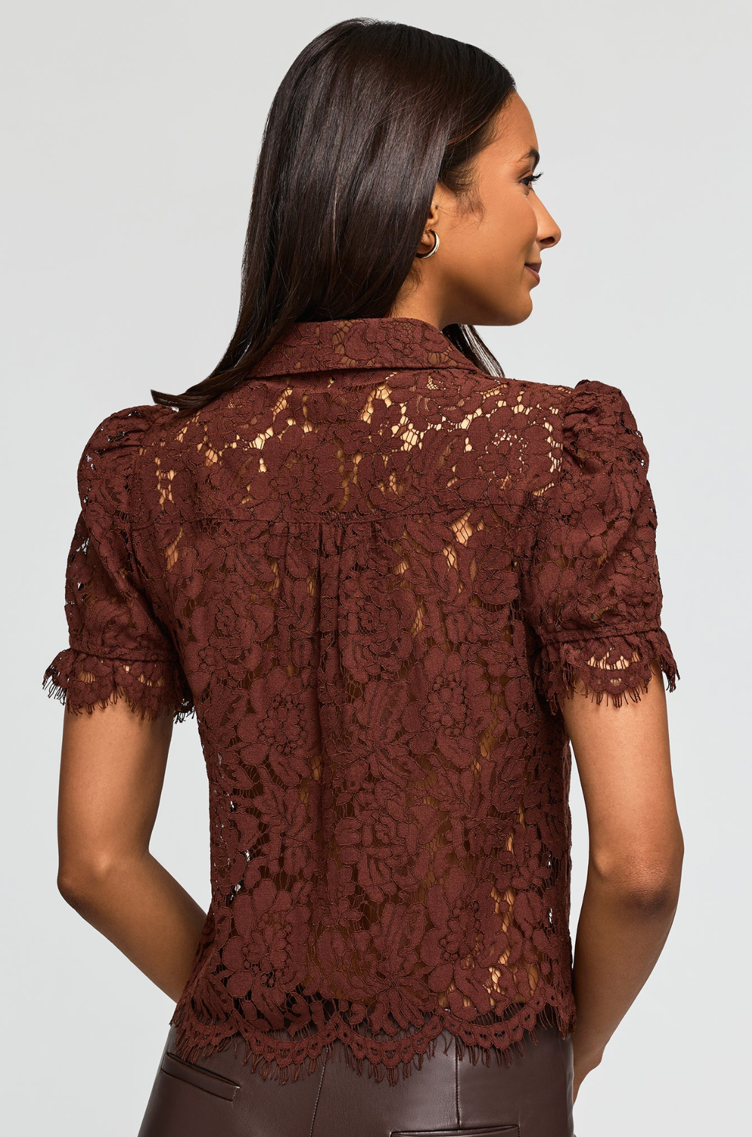 A woman shown from the back wearing a short-sleeve lace blouse in a deep burgundy brown color, paired with dark brown leather pants, against a light gray studio background.