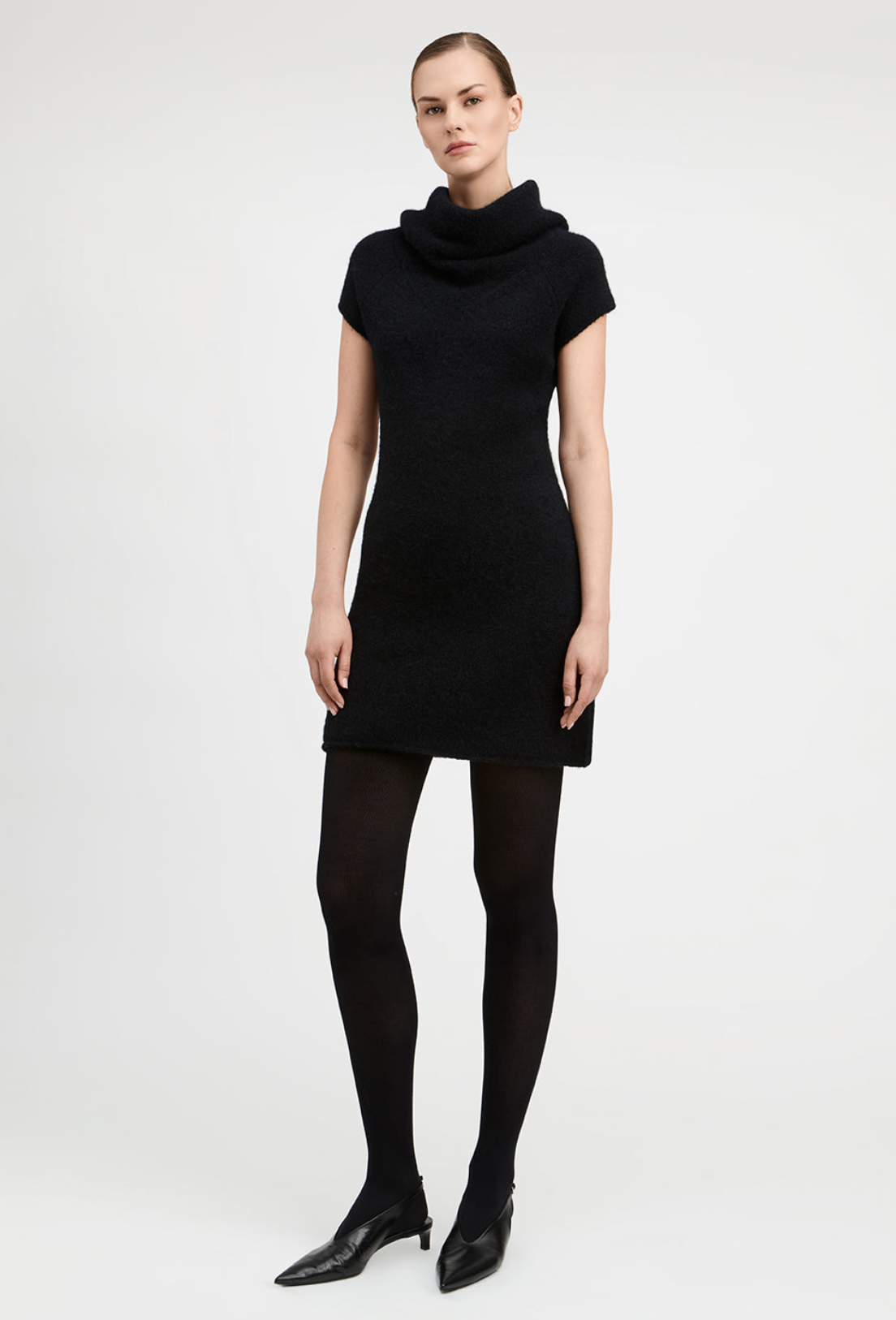 A model wearing a black short-sleeve cowl-neck knit dress, black tights, and black pointed-toe ankle boots, standing against a light gray studio background.