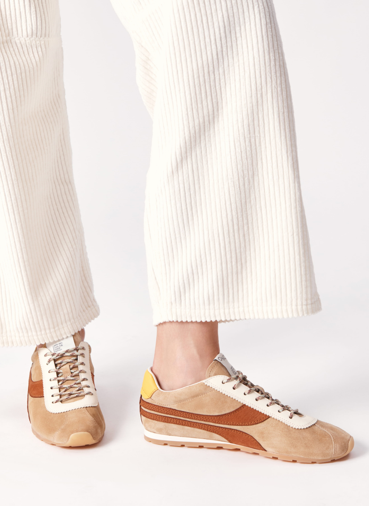 A low-top suede sneaker in beige and brown with a white leather side stripe, beige laces, a yellow heel tab, and a gum sole, shown in profile against a white background.