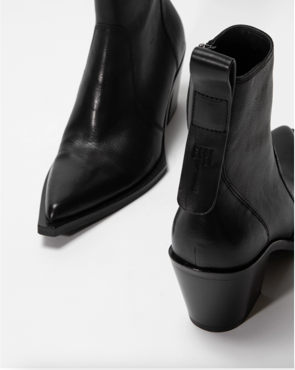 A pair of black leather ankle boots with pointed toes and low block heels, shown on a clean white studio background. One boot is positioned to show the side profile with a pull tab, and the other shows the pointed toe from a three-quarter angle.