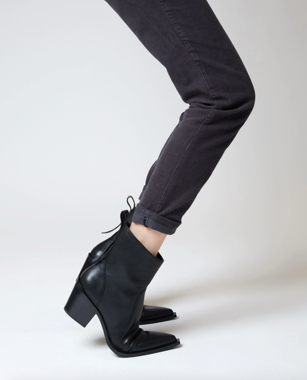 Black leather ankle boots with a block heel and pointed toe, shown on a model wearing dark gray cuffed jeans against a light gray studio background.