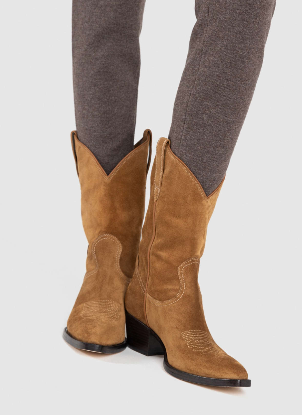 A pair of tan suede cowboy boots worn with gray pants, shown from the knees down against a light gray studio background.