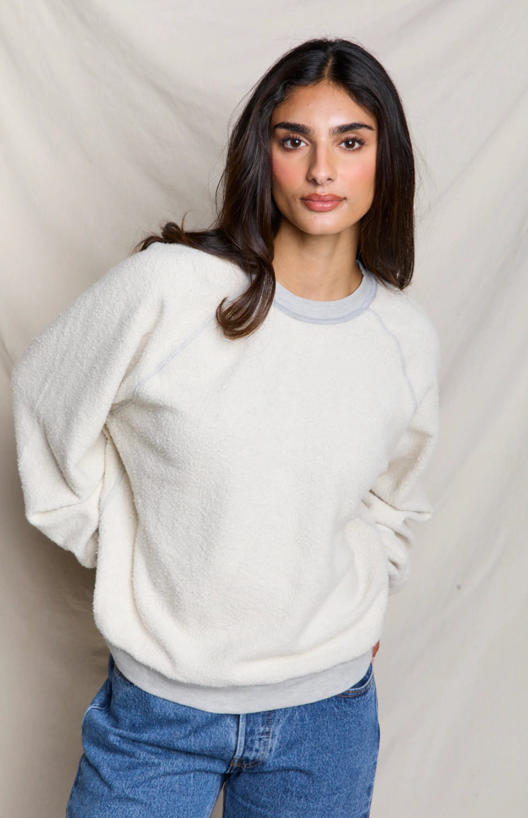 A woman wearing a cream-colored, textured fleece crewneck sweatshirt with light gray trim on the collar and cuffs, paired with blue jeans, standing against a draped beige fabric backdrop.