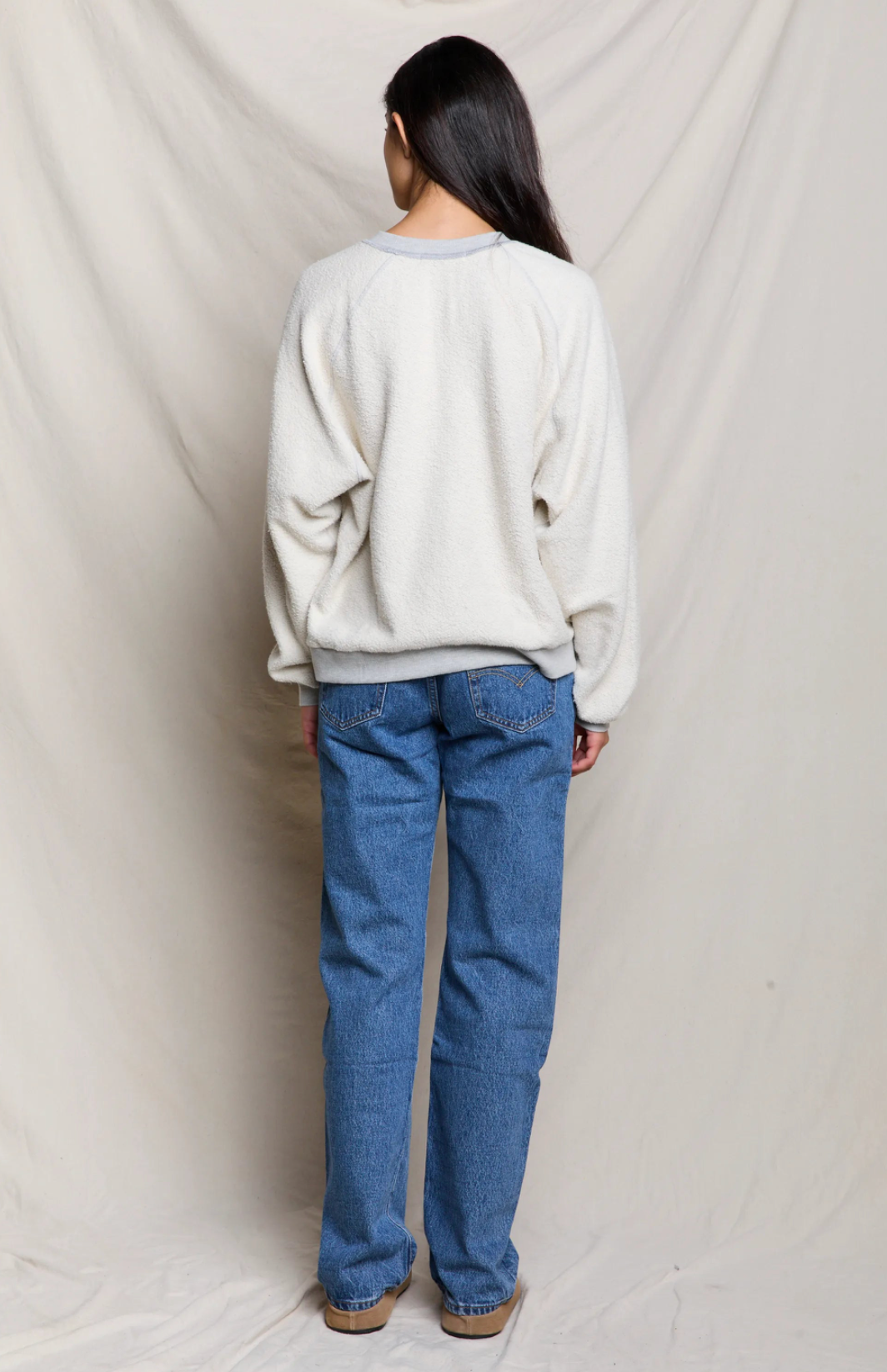 A woman is shown from behind wearing a cream-colored textured crewneck sweatshirt and blue jeans, standing against a draped off-white fabric backdrop.