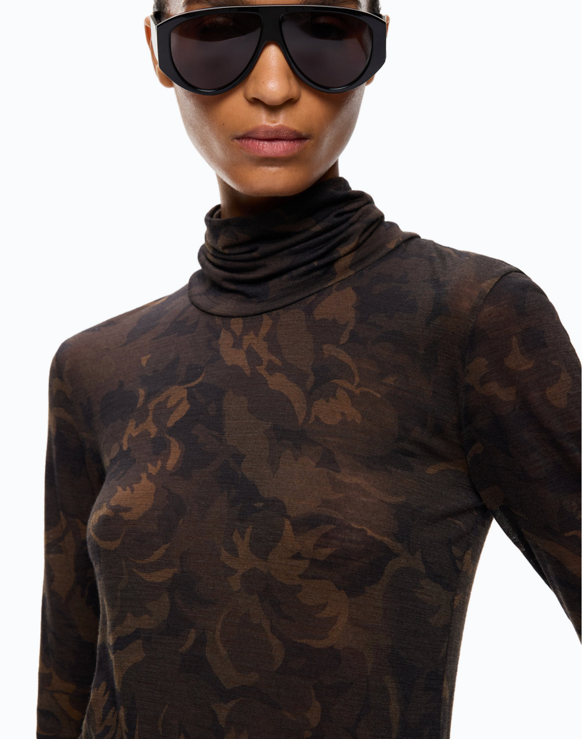 A close-up of a model wearing a sheer long-sleeve turtleneck top with a dark brown and black floral camouflage pattern, paired with oversized black sunglasses against a plain white background.